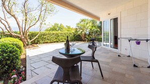 Une terrasse extérieure avec une table ronde en verre et des chaises en osier, une bouteille de vin et un verre.