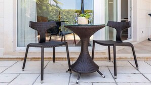 Une terrasse fermée par des baies vitrées, avec des meubles en osier, une table et une bouteille de vin.