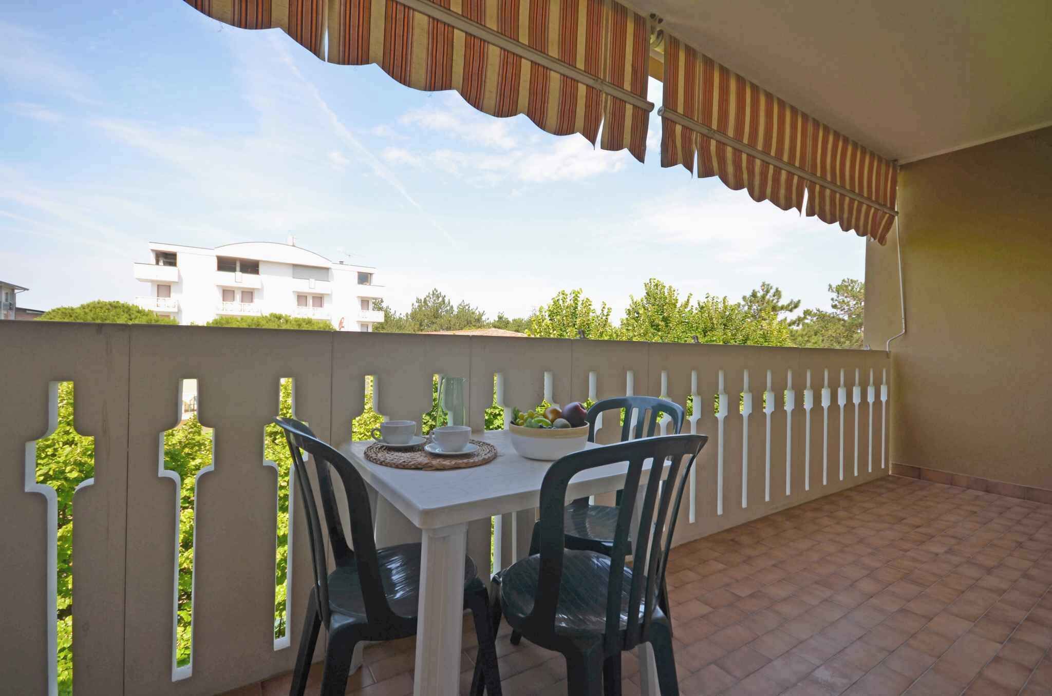 Uma varanda com mesa e cadeiras brancas, um toldo listrado e vista para um edifício e árvores.