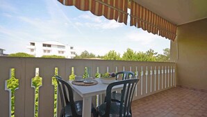 A balcony with a white table and chairs, a striped awning, and a view of a building and trees. - Holiday apartment with air conditioning and balcony (Bibione)