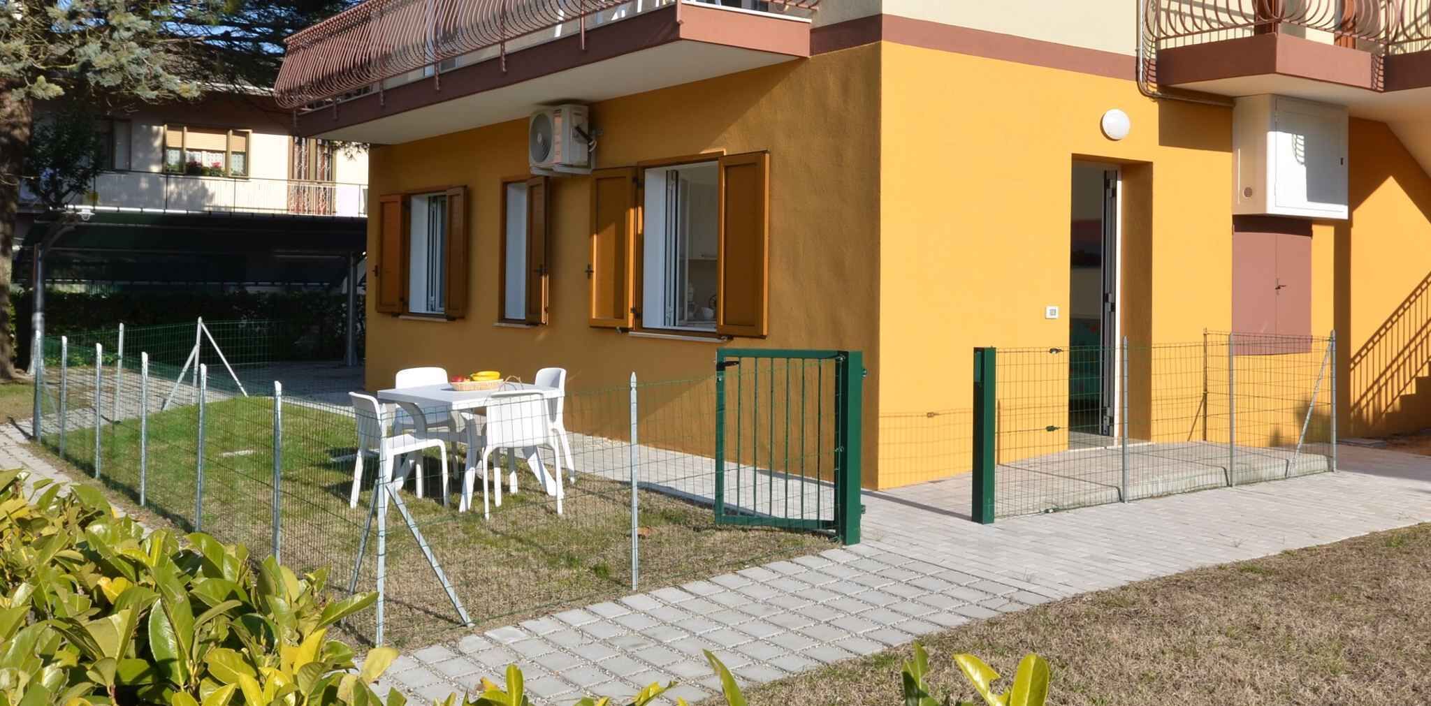 Un edificio amarillo con balcón, un jardín cercado con mesa y sillas, y una verja verde.