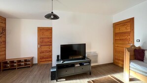 A room with a TV on a black stand, wooden doors, a bench, and a bed with a wooden headboard. - En toute amitié    . (Faucon-de-Barcelonnette)