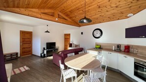 A modern kitchen with a wooden ceiling, a dining area with a round table and white chairs, a living area with a sofa, and a TV. - En toute amitié    . (Faucon-de-Barcelonnette)
