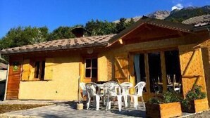 A small building with a tiled roof, a yellow exterior, and a patio with white plastic chairs. - En toute amitié    . (Faucon-de-Barcelonnette)