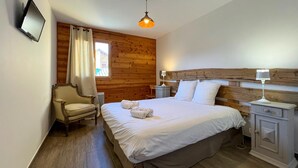 A bedroom with a wooden wall, a bed with white linens, two bedside tables with lamps, a chair, and a window with curtains. - En toute amitié    . (Faucon-de-Barcelonnette)