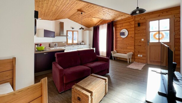 A cozy living room with a wooden ceiling, a red sofa, a wooden chest, and a kitchen in the background. - En toute amitié    . (Faucon-de-Barcelonnette)