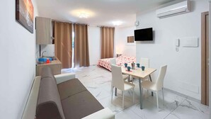 A modern hotel room with a bed, a dining area with a table and chairs, a kitchenette, and a TV mounted on the wall.