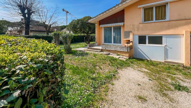 Une maison à deux étages avec un jardin, un chemin de gravier et un grand arbre en arrière-plan.