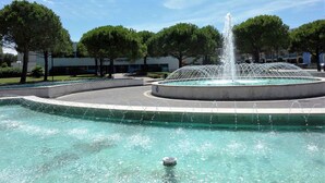 A fountain with water jets in a circular pool, surrounded by trees and a building with a sign that reads "THERMAL".