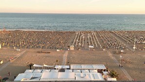 En strand med rader av parasoller och solstolar, fri sikt över havet och några få personer som promenerar längs stranden.