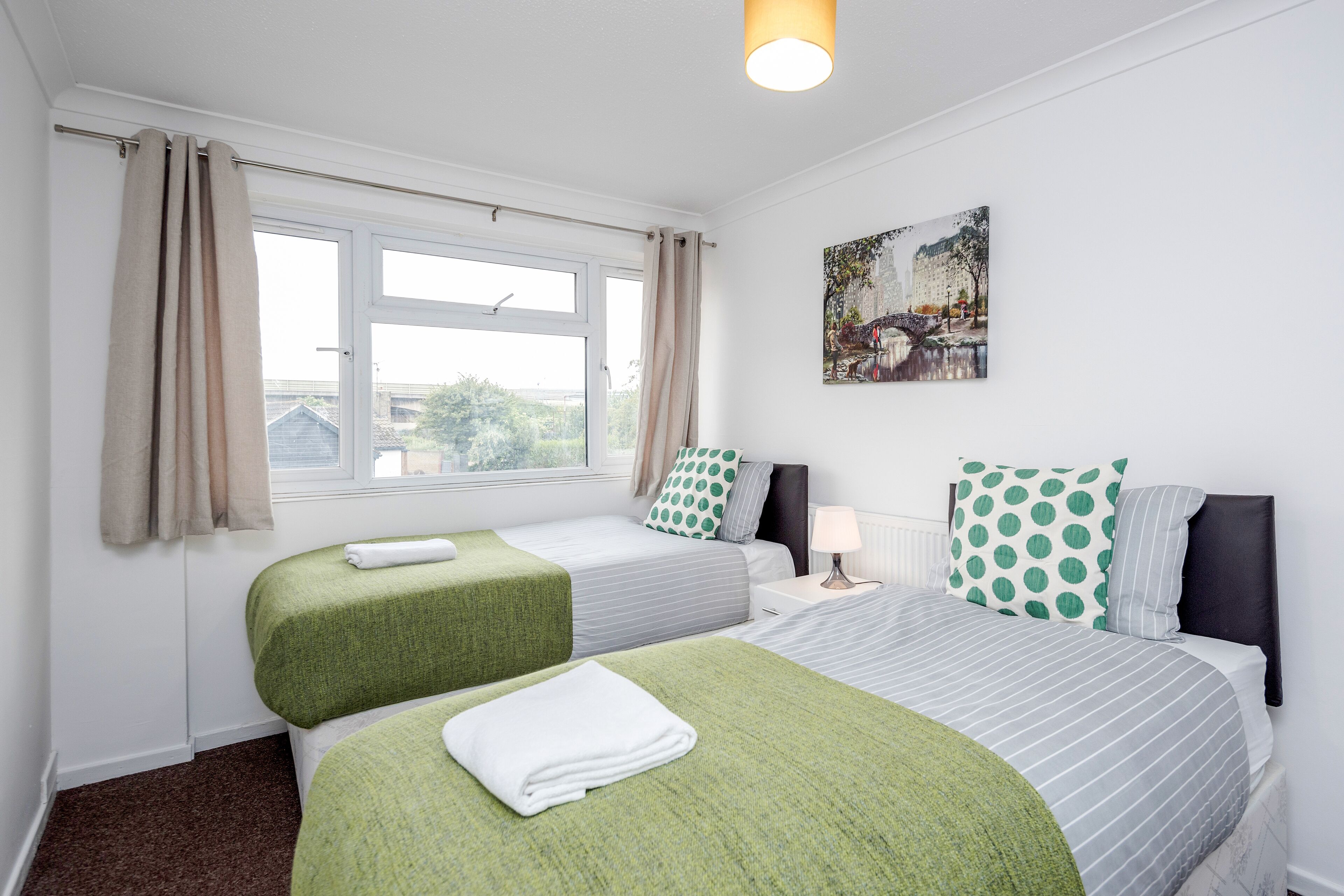 A hotel room with two beds, a window with curtains, a painting on the wall, and a bedside table with a lamp.