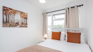 A bedroom with a bed, a nightstand, a lamp, a window with curtains, and a painting on the wall.