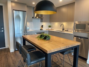 A modern kitchen with a wooden table, black chairs, a black pendant light, stainless steel appliances, and a potted plant.