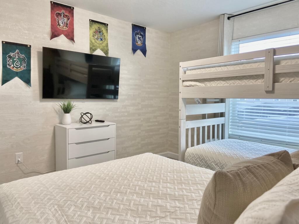 Une chambre avec un lit superposé, un téléviseur à écran plat, une commode et une fenêtre avec des stores.