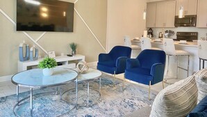 A modern living room with a TV, blue chairs, a round coffee table, and a kitchen in the background. - Beautiful 4BD/3BA Townhome with Pool 280 (Kissimmee)