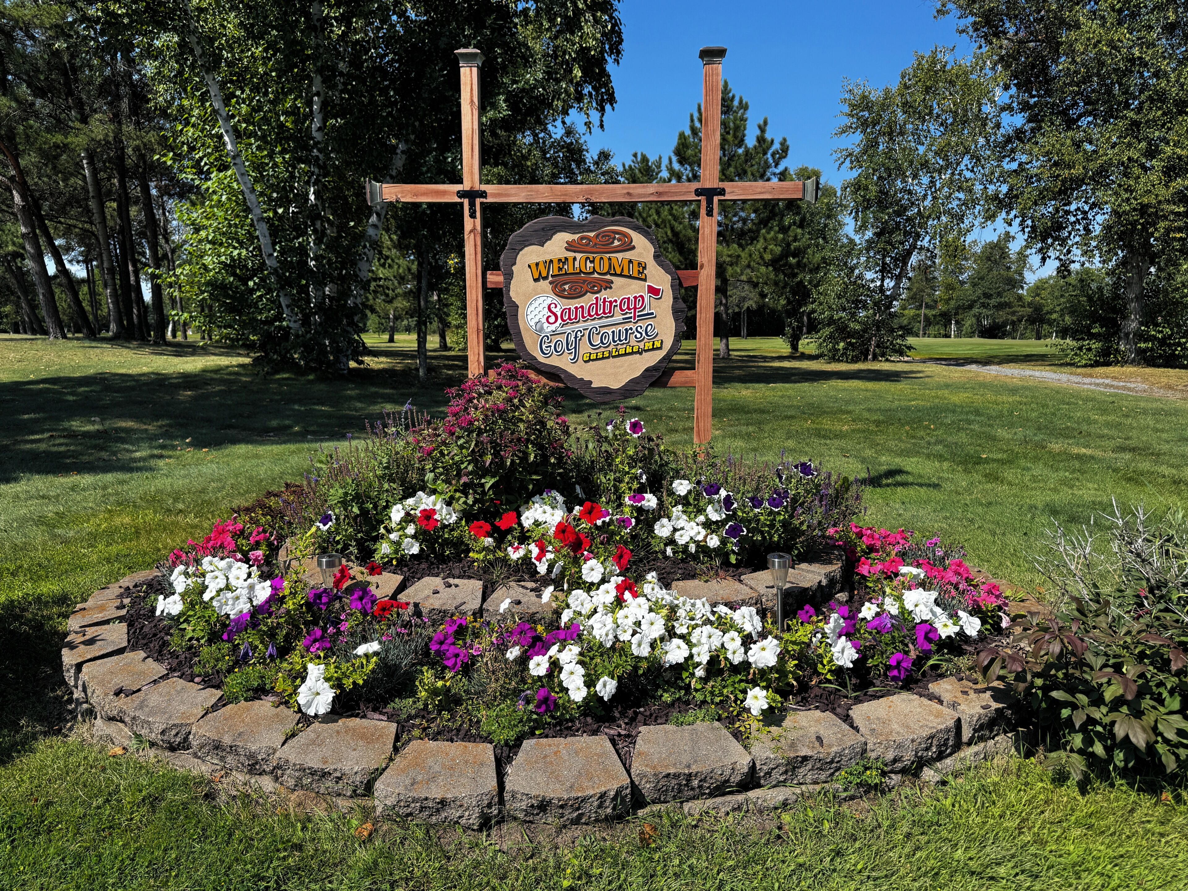 A welcome sign for Sandtrap Golf Course in Lakeville, MN, surrounded by a flower bed with various flowers.