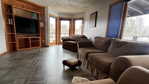 A living room with a large TV, wooden shelving, a sofa, and a patterned floor. - Large Private Condo on the Golf Course close to Many Lakes! (Cass Lake)