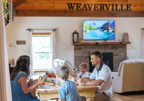 Una familia cenando en una habitación con chimenea, un televisor montado en la pared y un cartel que dice 'WEAVERVILLE'.