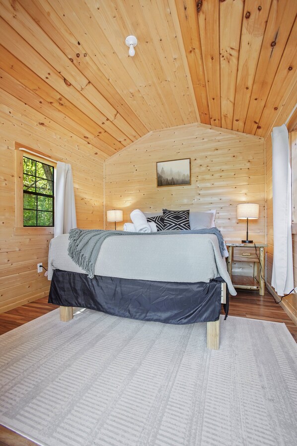 A wooden cabin bedroom with a bed, bedside lamps, a window with curtains, and a painting on the wall.