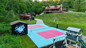 A tennis court with a 'GAME ON!' sign, surrounded by a wooded area and a lodge.