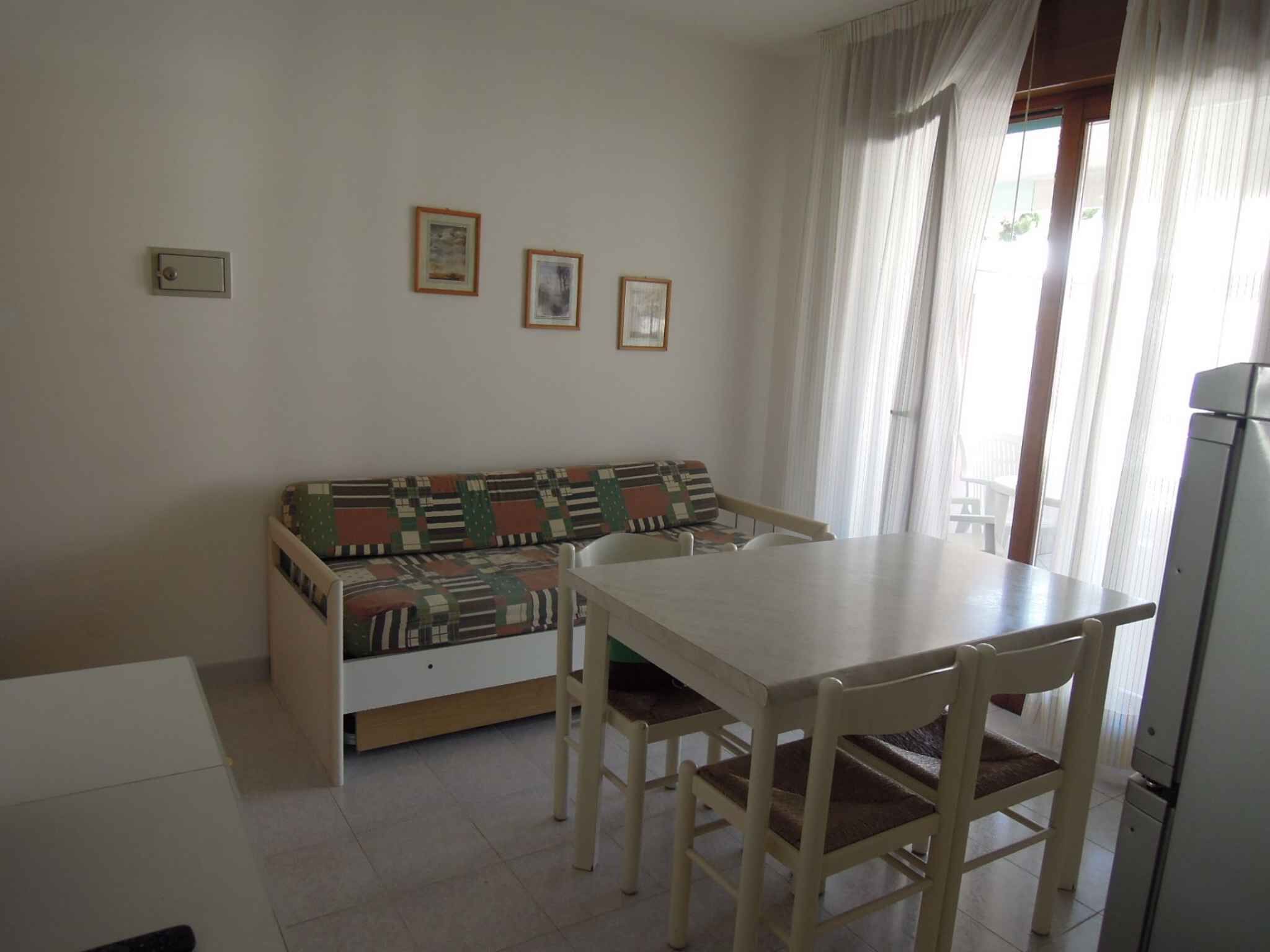 Een kleine, schone kamer met een bank, een tafel en stoelen. Er hangen drie ingelijste foto’s aan de muur.
