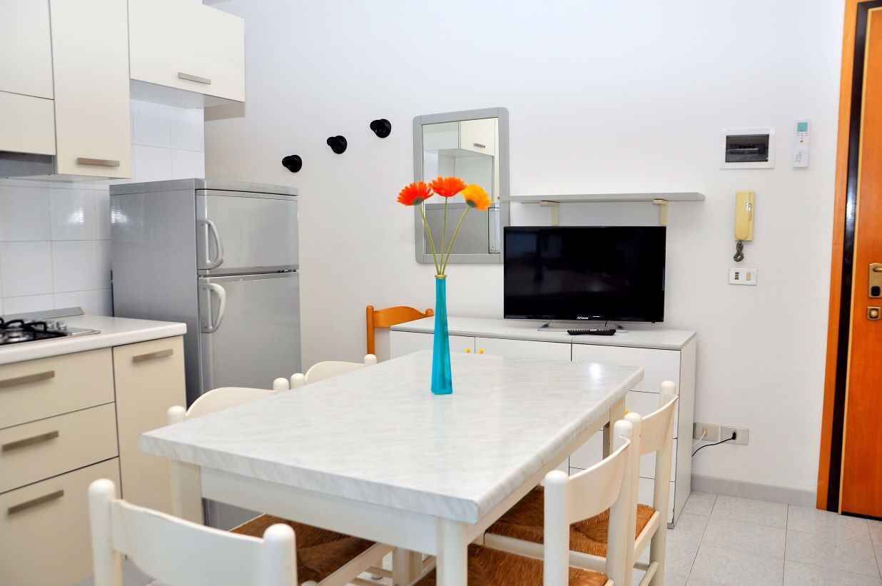 A compact kitchen with white cabinets, a stainless steel refrigerator, a dining table with white chairs, a vase with flowers, a flat-screen TV, and a wooden door.