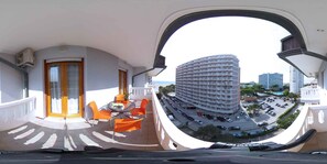 A balcony with a view of a multi-story building, a street with parked cars, and a clear sky.