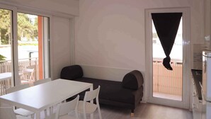 A compact room with a white table and chairs, a dark sofa, and a sliding glass door leading to an outdoor area. - Holiday flat with air conditioning and balcony (Lignano Sabbiadoro)