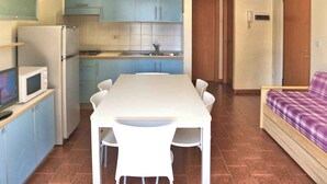 Een kleine keuken met blauwe kastjes, een witte tafel en stoelen. Er is een magnetron op een plank, een koelkast en een wastafel met een kraan. Een houten deur leidt naar een andere kamer.