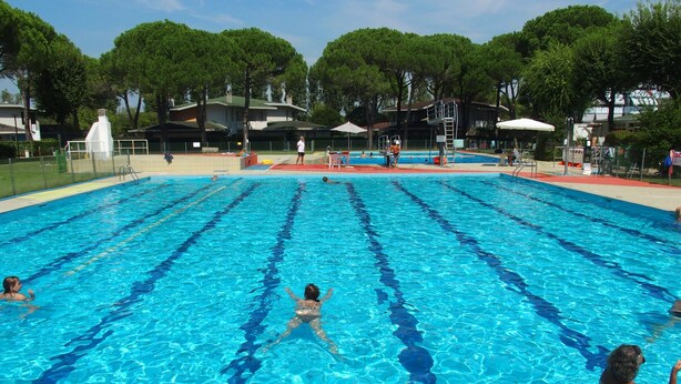 A swimming pool with clear blue water, surrounded by trees and buildings. - Holiday apartment with air conditioning, balcony and pool (Bibione)