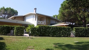 A single-story house with a flat roof, a chimney, and a front yard with a hedge and a lawn. - Holiday apartment with air conditioning, balcony and pool (Bibione)