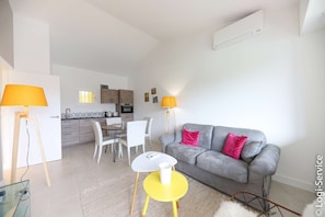 A modern living room with a grey sofa, yellow table, and white chairs. There is a kitchen in the background with wooden cabinets and a window.