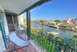 A balcony with a chair, a table, and a view of a river with boats and buildings in the background.