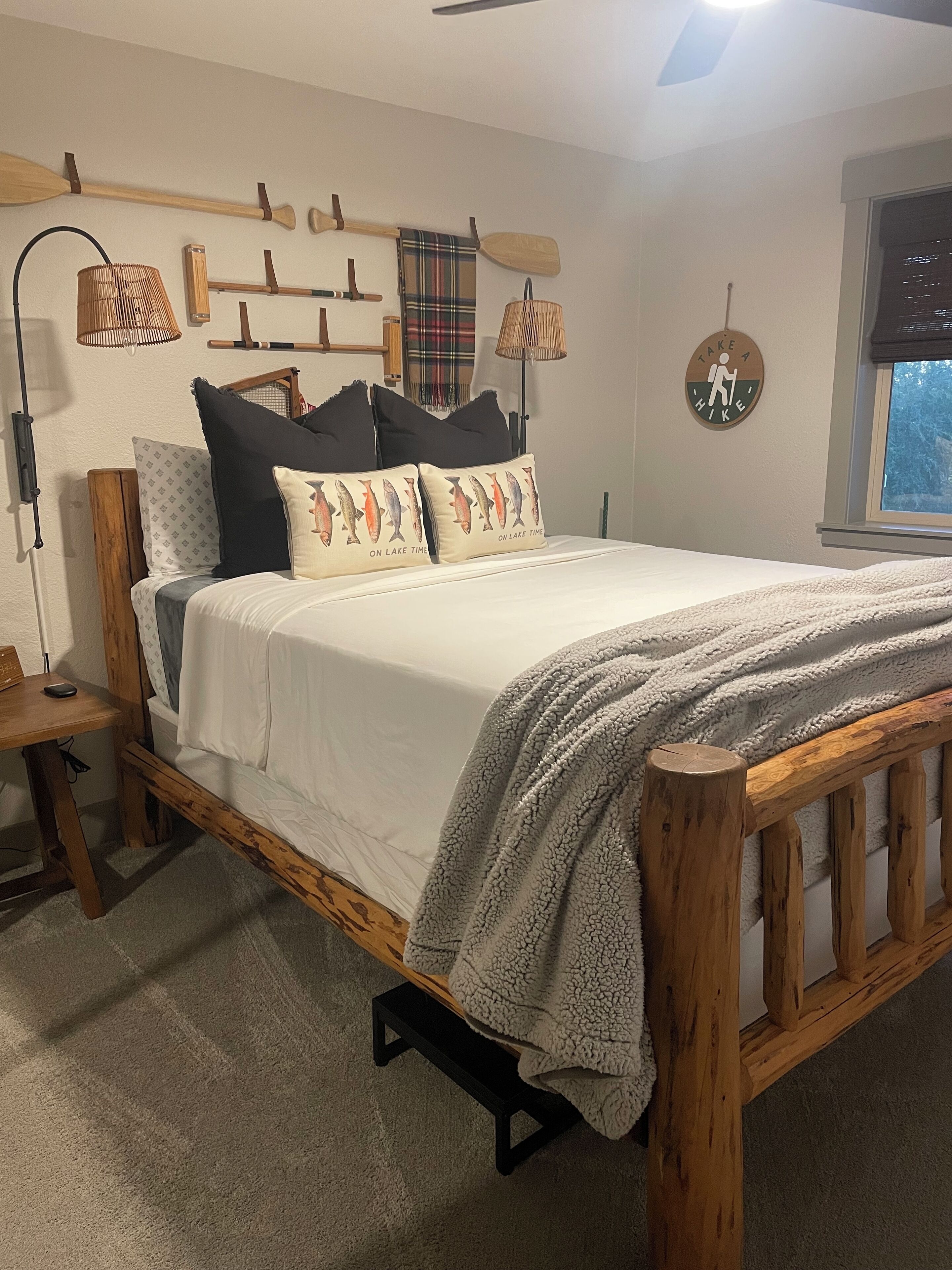 A cozy bedroom with a wooden bed frame, a large window with a view, and a wall-mounted shelf with paddles and a lamp.