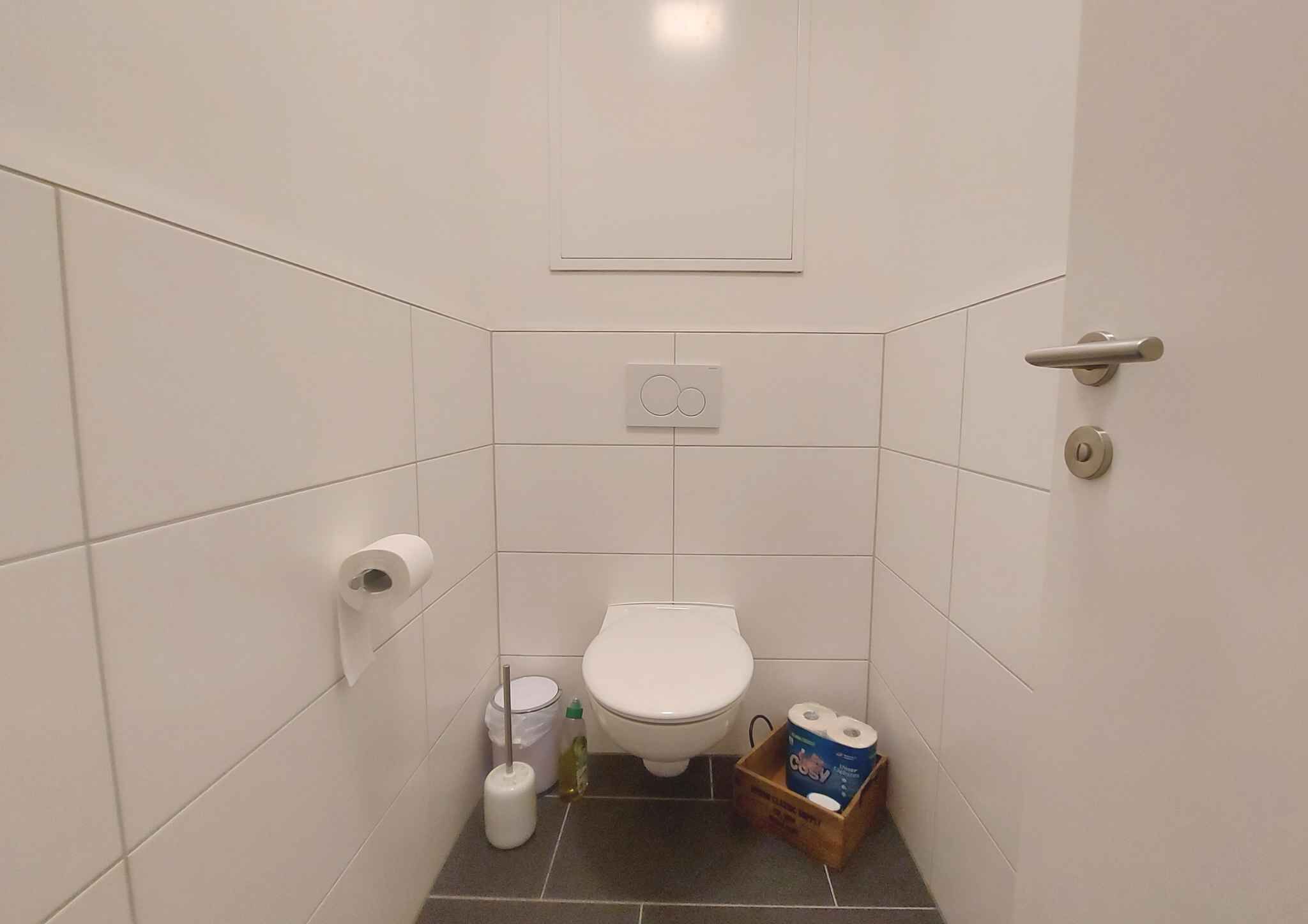Un baño pequeño y limpio con baldosas blancas, un inodoro, un secador de manos y un cubo de basura.