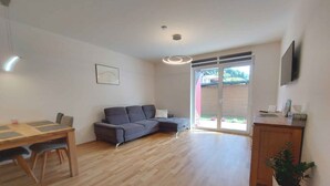 A modern living room with a wooden floor, a grey sofa, a dining table with chairs, and a sliding glass door leading to a backyard. - Holiday apartment new and cozy (Öblarn)