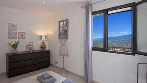 A modern bedroom with a large window, a dark dresser, a lamp, and a view of a cityscape. - Casa Bianca (L'Île-Rousse)