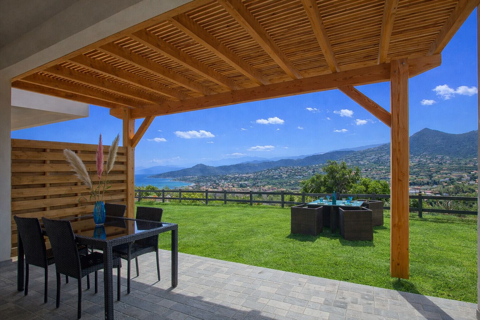 Un espace extérieur aménagé, abrité, avec une table et des chaises, un vase contenant des plumes d’herbe des pampas, et une vue sur une ville côtière et des montagnes.
