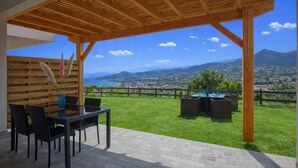A covered outdoor seating area with a table and chairs, a vase with pampas grass, and a view of a coastal town and mountains. - Casa Bianca (L'Île-Rousse)