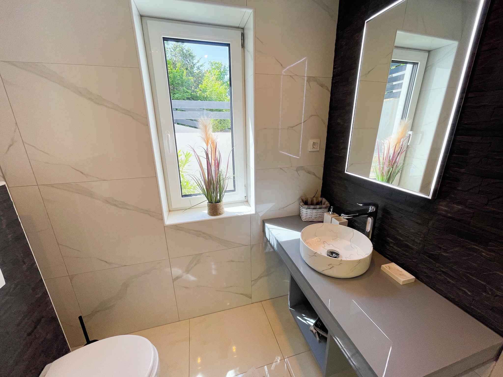 A modern bathroom with a large mirror, a white sink, and a window with plants.