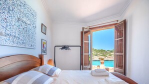 Une chambre avec un grand lit, une tête de lit en bois, une table de chevet, une lampe et une vue sur la mer par de grandes fenêtres ouvertes.