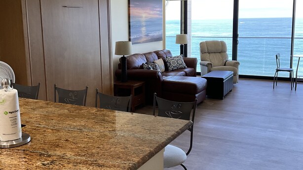 A modern kitchen with granite countertops, a dining area with a table and chairs, a sofa, and a view of the ocean through large windows. - floor to ceiling windows,180 degree view of ocean, 11th floor (San Diego)