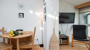 Un salon moderne avec une table ronde en bois, deux fauteuils en osier, un canapé noir, un téléviseur à écran plat et une grande fenêtre.