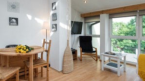 Un salon moderne avec une table à manger en bois, des chaises, une petite table d’appoint blanche et une grande fenêtre donnant sur la forêt.