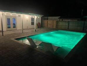 Una piscina rettangolare con bordo piastrellato, circondata da una recinzione in legno e un'area patio coperta con luci a stringa.