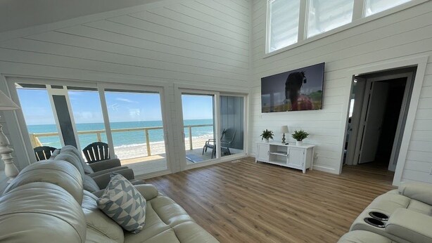 A spacious living room with a large beige sectional sofa, a wooden floor, and a large window overlooking a beach. - Seabatical - Oceanfront in Garden City Beach, SC (Garden City Beach)