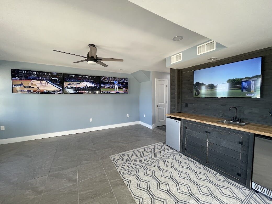 A spacious living room with a large flat-screen TV, a ceiling fan, and a kitchen area with a sink and cabinets.