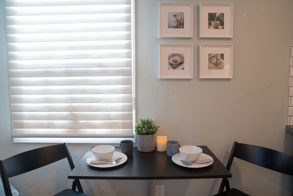 A dining area with a table set for two, a potted plant, and framed pictures on the wall.