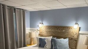 Quarto de hotel com uma cama grande, cortinas cinzentas, cabeceira decorativa, duas luminárias de cabeceira e uma pequena mesa amarela.