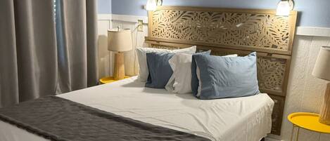 Een hotelkamer met een groot bed, grijze gordijnen, een decoratief hoofdbord, twee bedlampen en een kleine gele tafel.